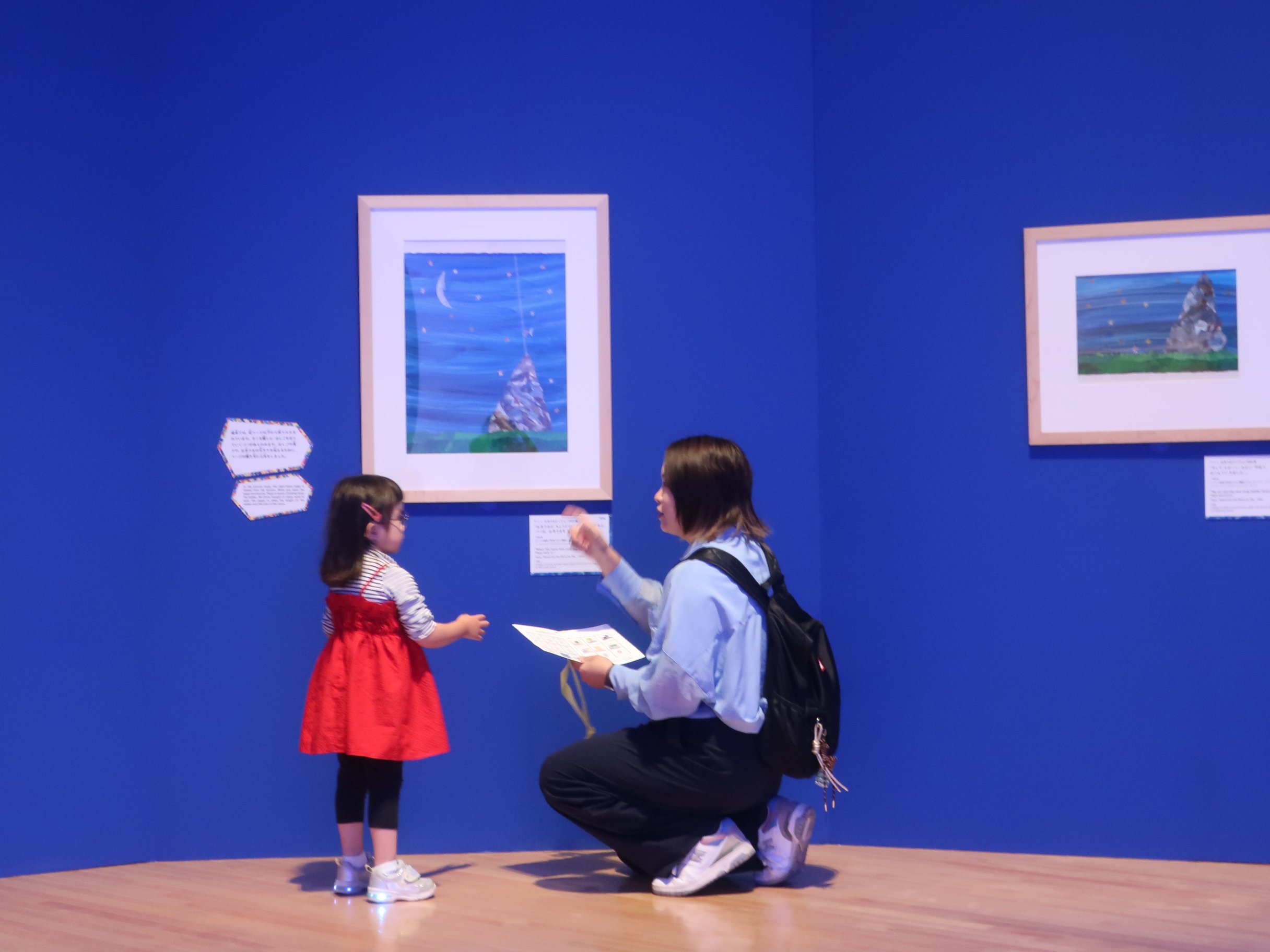 目の絵のある、青い壁の展示室。眼鏡をかけた赤いワンピースの小さな女の子と視線をあわせてしゃがんだ母親が、ワークシートを手に手話で話をしている。