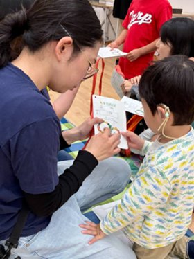 補聴器をつけた小さな男の子がスタッフのひざに手を置いて、ワークシートをさしだしている、スタッフは表紙にシールを貼ろうとする。