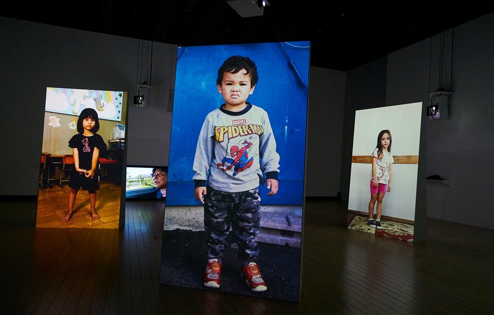 There are three projected screens and a small monitor on the floor in a dark gallery. The monitors show a middle-aged woman talking; the three screens show children, ranging from pre-school to early primary school age, one on each screen, looking at the viewer.