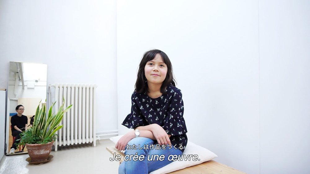 A girl of about 10 years old is sitting on a bench in a white room, facing us. There is a mirror at the far left of the room, in which an adult woman is reflected. The girl and the woman seem to be having a conversation. The subtitle says: 'I create an artwork'.