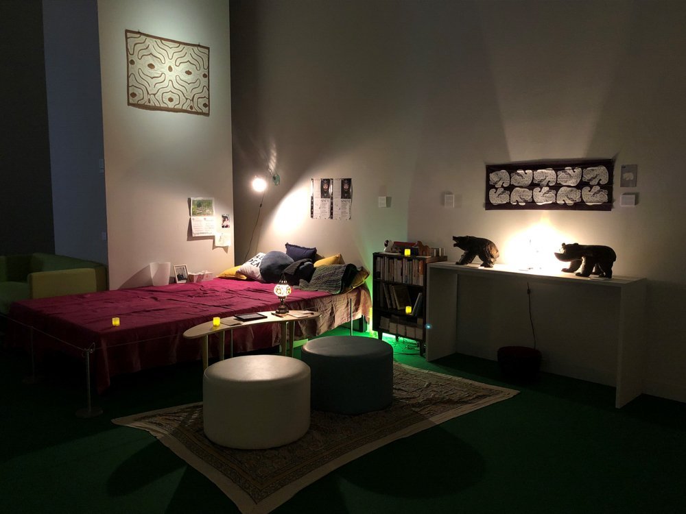 In the center of the dimly lit room is a bed covered with pink sheets. On the side is a bookshelf, a stand with a carved wooden bear figurine, a small table, and two round stools. A patchwork tapestry hangs at the top of the wall just next to the bed.