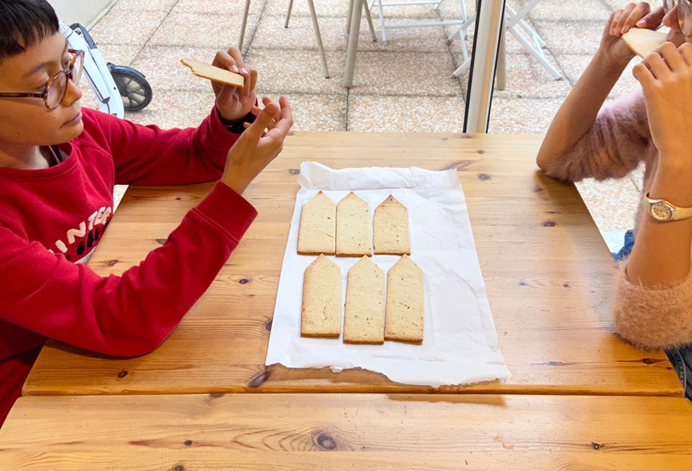 Two figures are facing each other, eating cookies at a café table. The cookies are plain dough, flat and have an elongated pentagonal shape, like a house or a pencil.