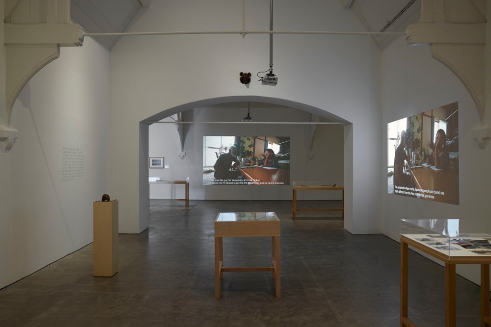 Installation view in a gallery in the form of several connected, white-walled rooms. A man and a woman are talking to each other in a projection on the wall at the back and right of the screen. There are several display cases in the gallery.