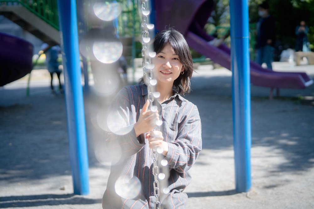 A park with strong contrasts of light and shadow. In the centre of the image, a swing chain reflects light like a sun catcher. On the far side of the light, there is a woman in her twenties holding the swing chain and smiling.