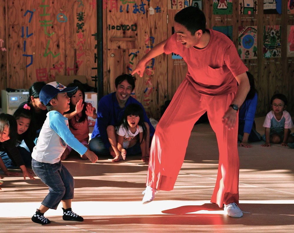 Preschool children and their parents sit in a circle. In the midst of the circle, a boy wearing a cap and a man in red T-shirt and loose-fitting trousers are dancing face to face.
