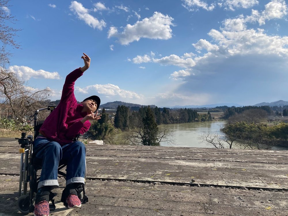 There is a blue sky with soft clouds in the upper half of the image, under which a river flows slowly. There is a man in a wheelchair dancing on what looks like a wooden deck on the riverbank, with his eyes closed and his hands raised wide.