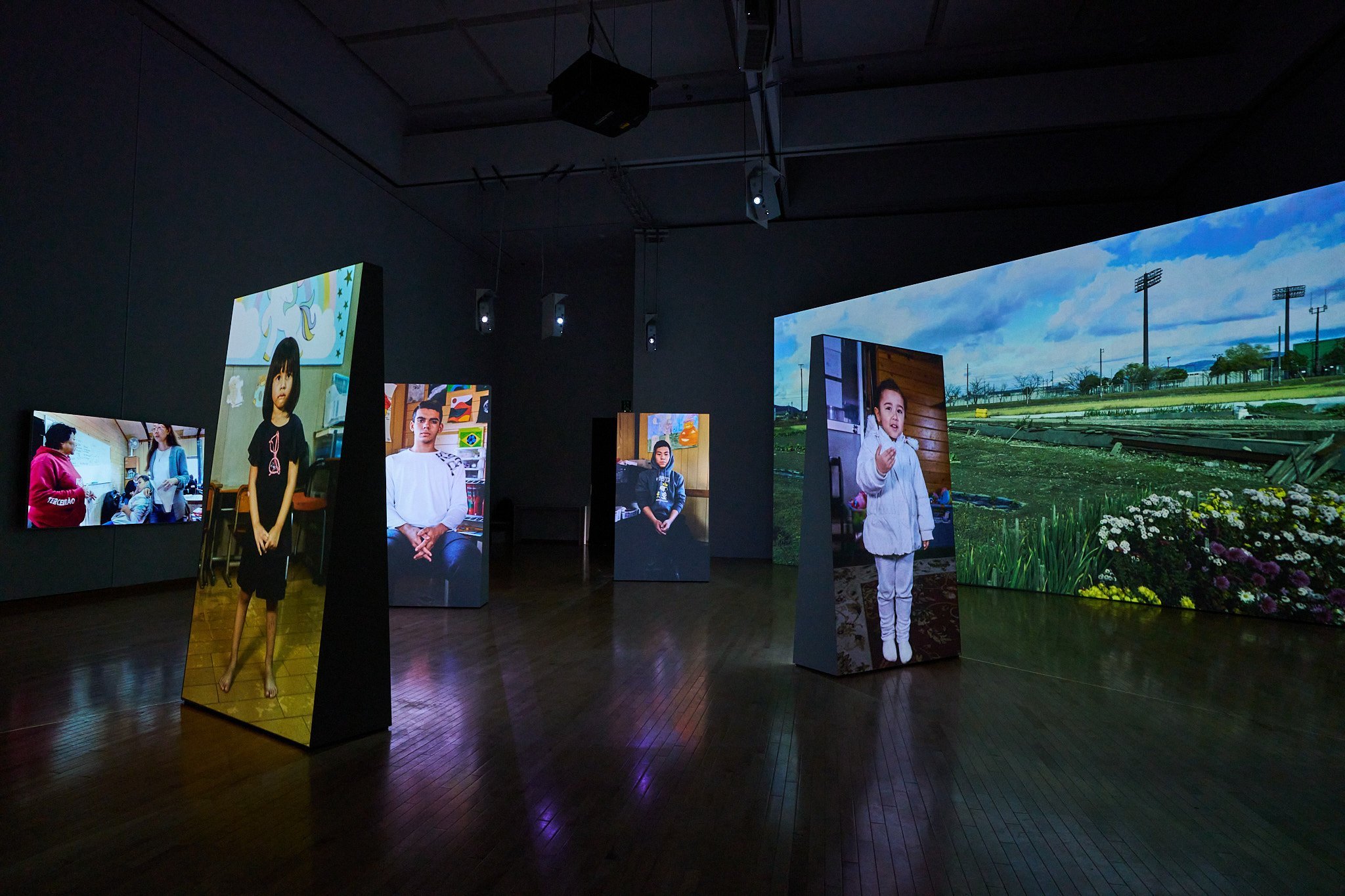 Six projected screens float in the darkened exhibition room. The largest horizontal screen on the right shows a plain, field-like area with no tall buildings. The horizontal screen on the left wall shows a woman in a pink hoodie and a tall woman in a light blue cardigan. Four vertical screens placed in the center of the room show one child each, ranging from toddler to high school age, looking at the viewer.