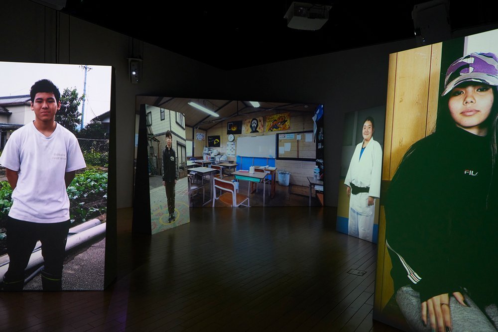 Five projected screens are shown in a dark gallery. At the far centre is a school classroom. On the four screens in the foreground, a boy and a girl of junior or senior high school age are shown, one on each screen, looking at the viewers.