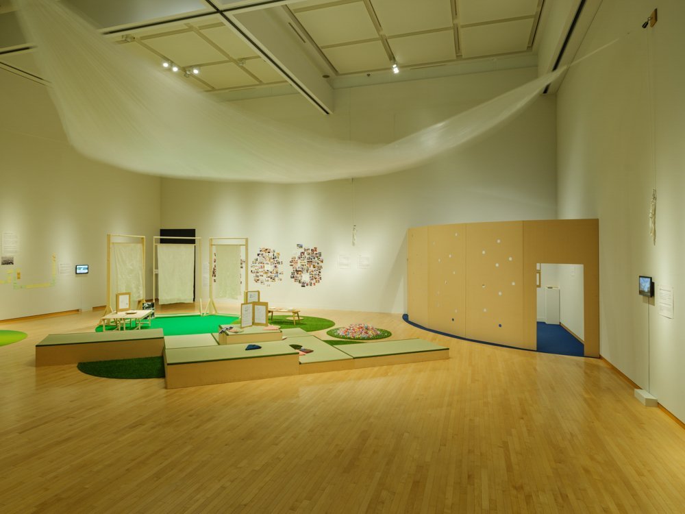 A view of the exhibition room with wooden floor and white walls. A large white curtain-like structure hangs from the ceiling and seems to rustle in the wind. In the center of the room, there is a small tatami-mat floor with several small cushion-like objects. At the far right is a fan-shaped space separated by a wall with peepholes. Beyond the tatami mats are three rows of wooden frames covered with what looks like white sheets.