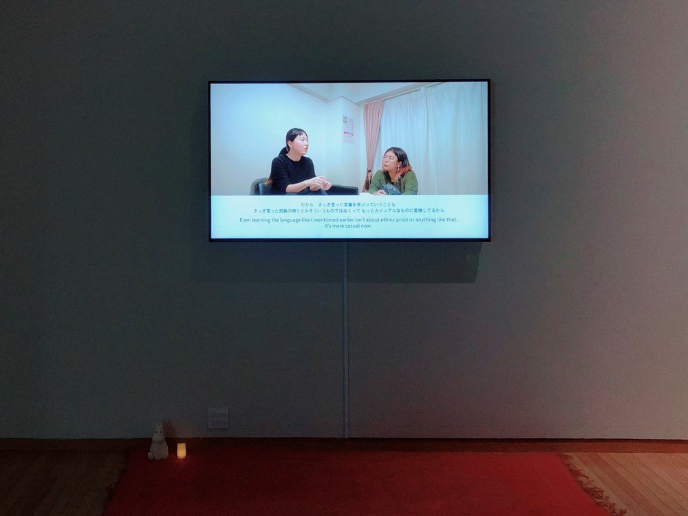 A monitor is attached to a white wall in a dark room. The image on the monitor shows two women talking in a dull meeting room. The woman on the left has her black hair tied in a single knot and is speaking while staring slightly upward. The subtitle says, 