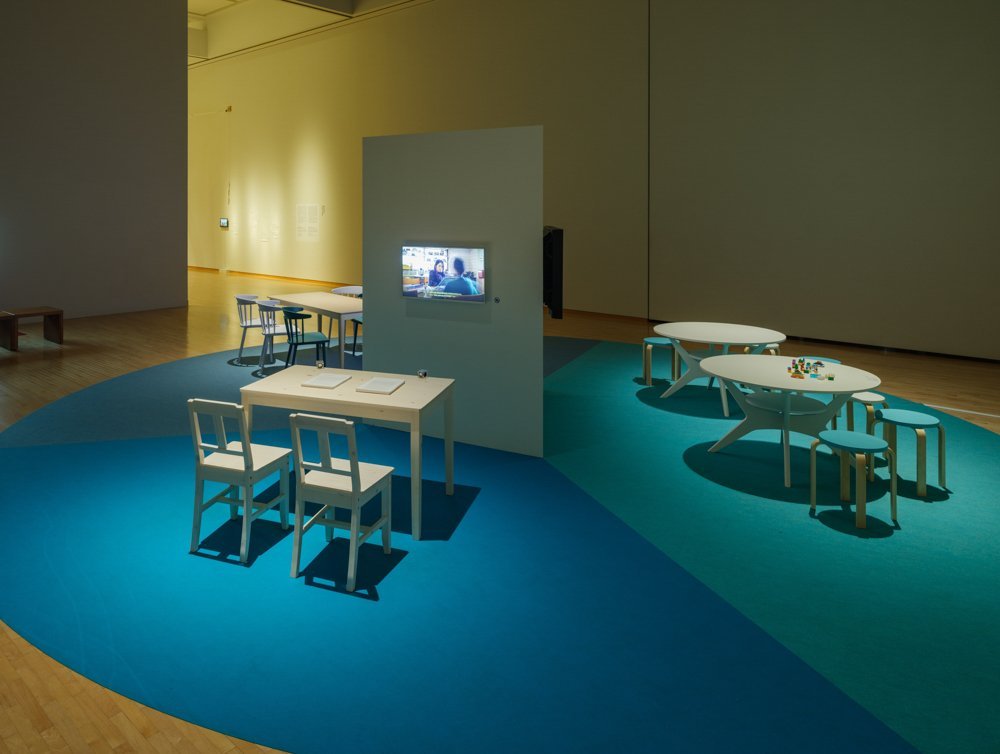 There is a carpet divided into three areas like a pie chart, with a white column with monitors in the center. The blue-carpeted area in the front has a small desk and two chairs, and the monitor shows two figures talking across a table. The light blue area on the right has a round-topped low tables and some round stools. The dark blue area on the far left has a long dining table and some back chairs.