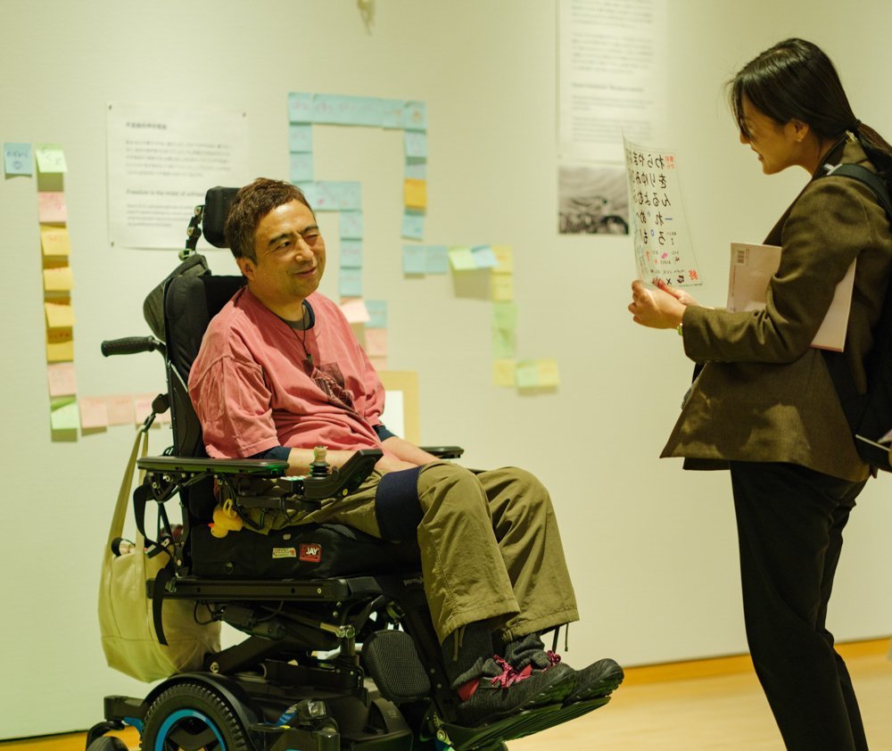 A man in a large electric wheelchair, wearing a T-shirt and cargo pants, smiles, closes one eye, and looks at an eye gaze communication board held by a woman standing across from him. The woman holding the board wears a green jacket and her long black hair is tied in a single knot. She bends her knees slightly to bring the board down to the man's eye level and smiles. On the wall behind them are a number of sticky notes.