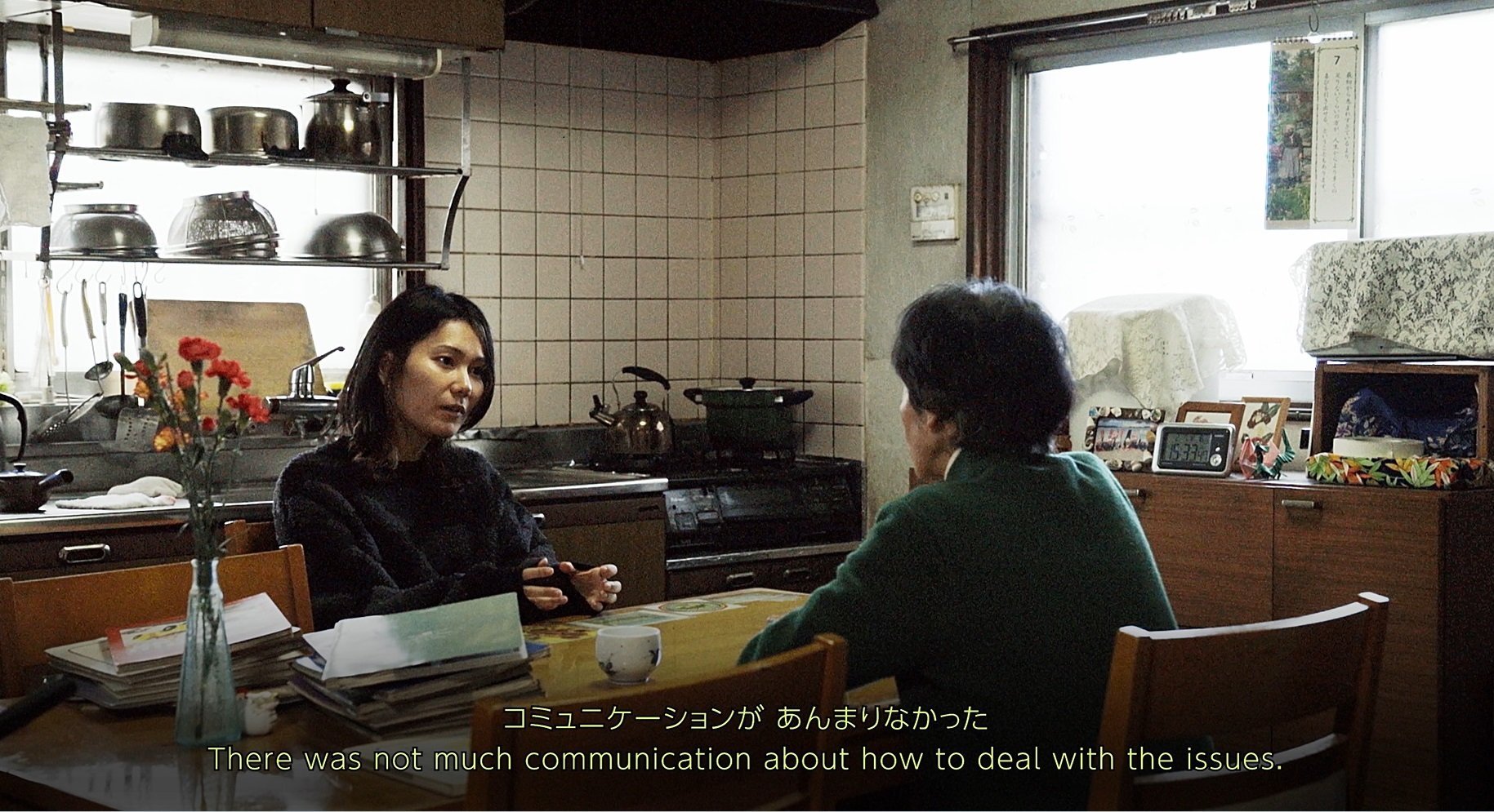 Two women are having a conversation at a kitchen table. On the table are some carnations and an old photo album. The young woman with her back to the well-worn kitchen has her body turned toward us, and the expression on the face of the older woman facing the young woman is not visible to the viewer. The young woman is speaking and the subtitle says, “These was not much communication about how to deal with the issues.”