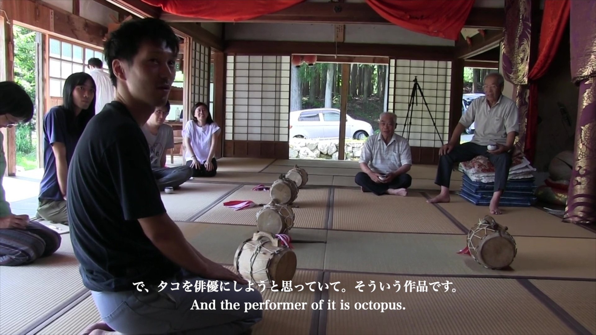 お寺や古民家のような畳敷きの部屋の中で、７人の人物が座っています。４人は正座をしており、2人は胡坐、そして高齢の男性は重ねた座布団をイスのようにして座っています。「で、タコを俳優にしようと思っていて。そういう作品です」と字幕が表示されており、手前の正座している男性が発言者のようです。