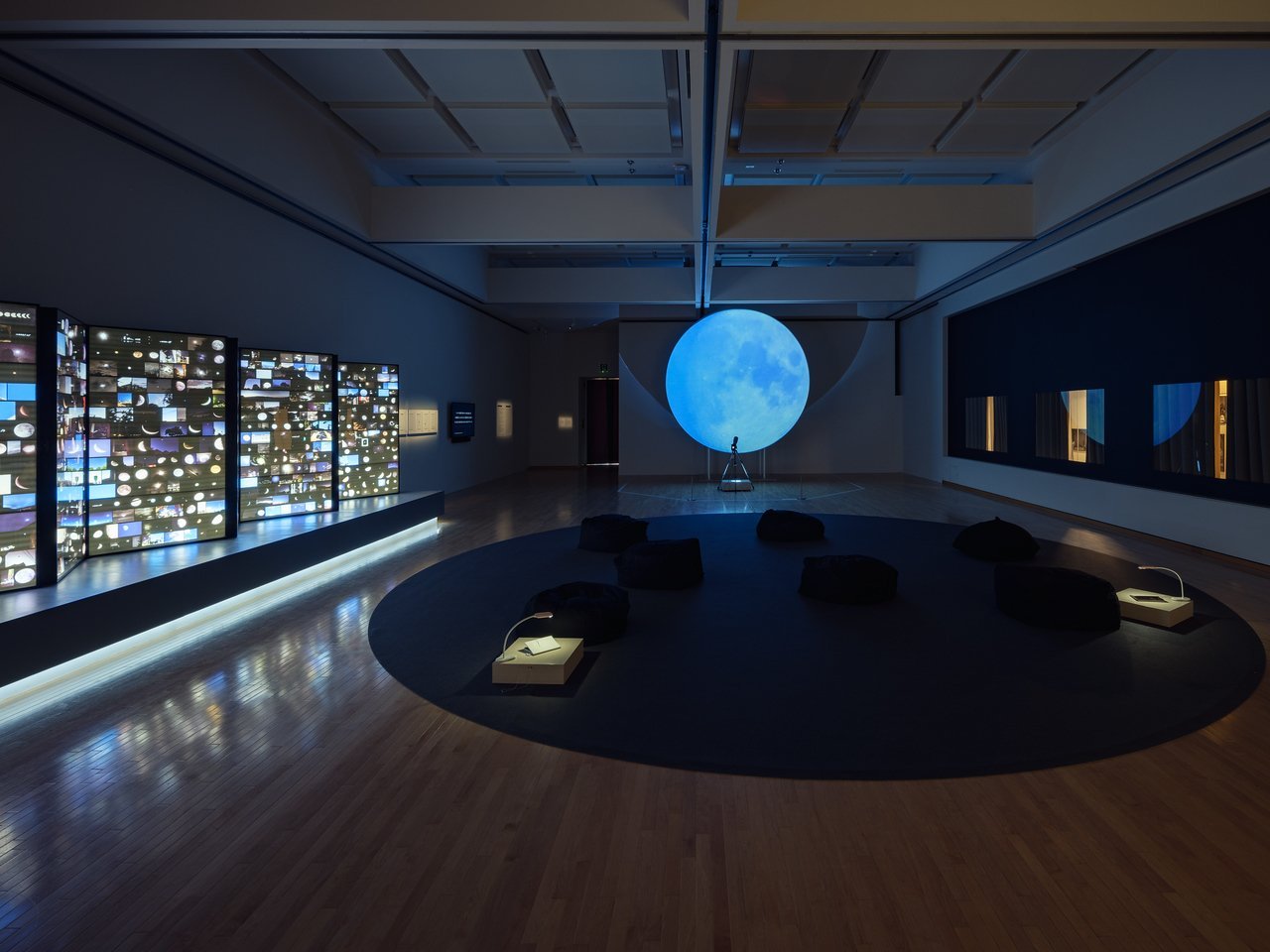 縦長の展示室の全体を見ている写真です。一番奥には、大きな青い月が浮かんでいます。その手前の床には、黒いカーペットが丸い形に敷かれています。右側の壁は、カーテンのついた窓のように見え、オレンジ色の光がカーテンの隙間から見えています。左側は青い光が印象的で、小さい写真がたくさん貼りこまれたライトボックスのようなものが並んでいます。